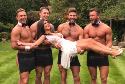 Four male butlers carry a laughing bride during a hen party in a garden.